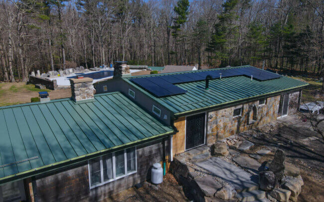 Aerial view of a building with a green metal roof and solar panels, surrounded by a forested area with rocks and moss.