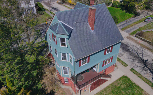 Aerial view of a residential neighborhood with a large, multi-story house with a complex roof structure and architectural details, surrounded by other houses and greenery.
