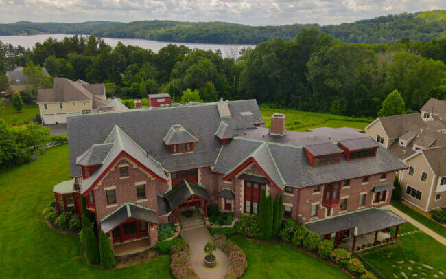 Aerial view of the rooftop and exterior of a large, multi-story red brick building with a complex roof structure and numerous dormers. The building is surrounded by green trees and landscaping, with other buildings and homes visible in the background.
