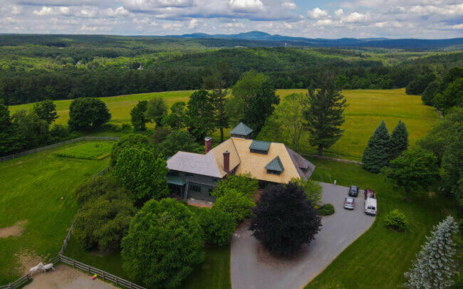 An aerial view of a large, multi-level building with a complex roofing structure featuring a mix of yellow brick and green metal panels. The building is situated in a picturesque, rural setting with rolling hills and forests in the background.