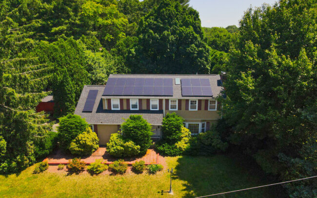 An aerial view of a large, two-story house with a gray roof and siding, surrounded by a well-maintained lawn and mature trees.