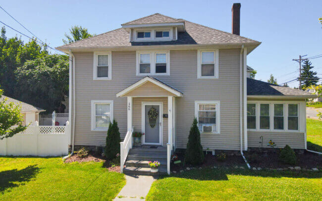 An aerial view of a large, two-story house with a gray roof and siding, surrounded by a well-maintained lawn and mature trees in a residential neighborhood.