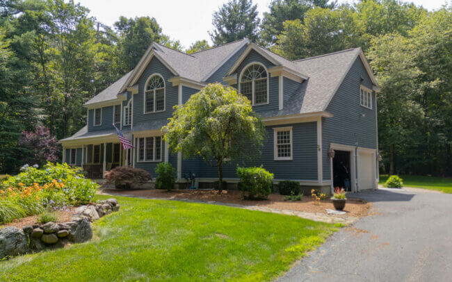 A large two-story house with a gray roof, yellow siding, and arched windows, surrounded by lush greenery and landscaping.