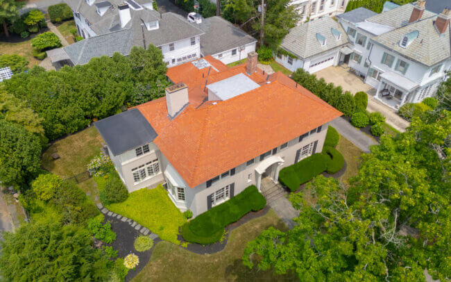 An aerial view of a large, two-story house with a red tile roof, surrounded by trees and a grassy yard.