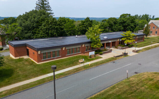 Aerial view of a multi-story brick building with a flat roof surrounded by trees and a winding road.