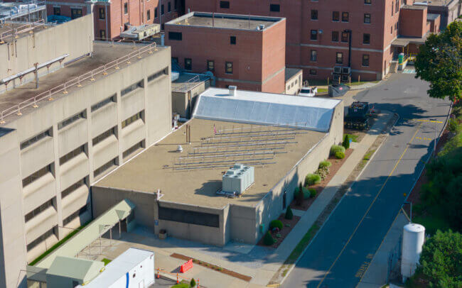 Aerial view of a multi-story brick building with a complex roof structure surrounded by trees and a parking lot.